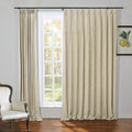 A living room scene with light beige linen pinch pleat curtains on a black rod framing a window with white grilles overlooking a sunny outdoor area, a white antique-style armchair with studded trim, and a framed botanical lemon print on the wall