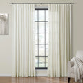 Pleated cream-colored curtains flank a black-gridded window in a modern living room, alongside a monochrome tree painting, a beige armchair with a textured throw, and a warm hardwood floor