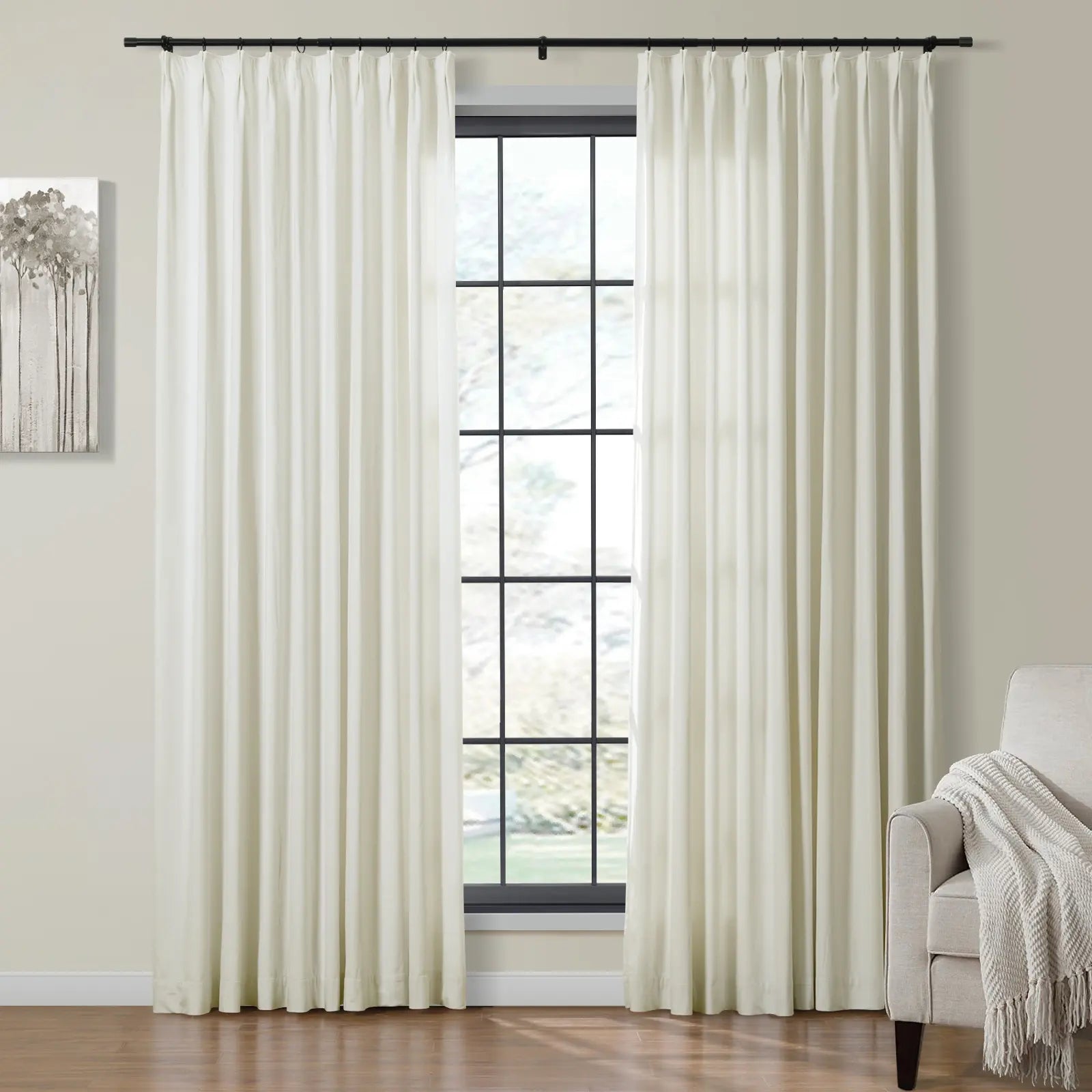 Pleated cream-colored curtains flank a black-gridded window in a modern living room, alongside a monochrome tree painting, a beige armchair with a textured throw, and a warm hardwood floor