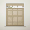 Natural light beige top-down bottom-up woven wood blind installed in a white window frame with a brass ring pull partially revealing green foliage and subtle pane shadows