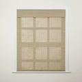 Photograph of a window in an interior room featuring a light beige woven Roman blind with visible horizontal folds and faint grid-like shadows cast upon it from behind, framed by white molding