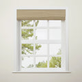 Photograph of a white multi-pane window featuring a tan woven Roman blind, set against an off-white wall with blurry green trees visible outside