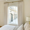 A bright bedroom interior with an arched window, cream drapes on a gold rod, and plush white quilted bedding, revealing an exterior view of a brick building with arched garage doors, decorative wall lanterns, and snow-covered ground