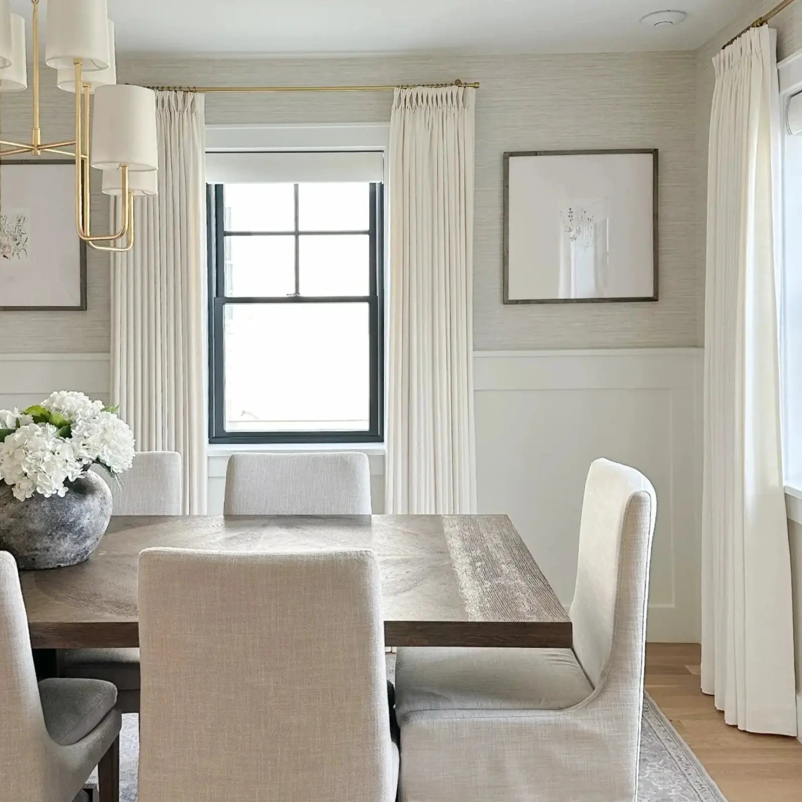 Neutral contemporary dining room featuring a dark wood dining table, light upholstered chairs, a grey vase with white hydrangeas, and a modern gold chandelier against textured wallpaper