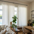 Interior view of a modern living dining room featuring sheer white linen curtains a large fiddle-leaf fig plant a geometric black chandelier and a visible outdoor swimming pool