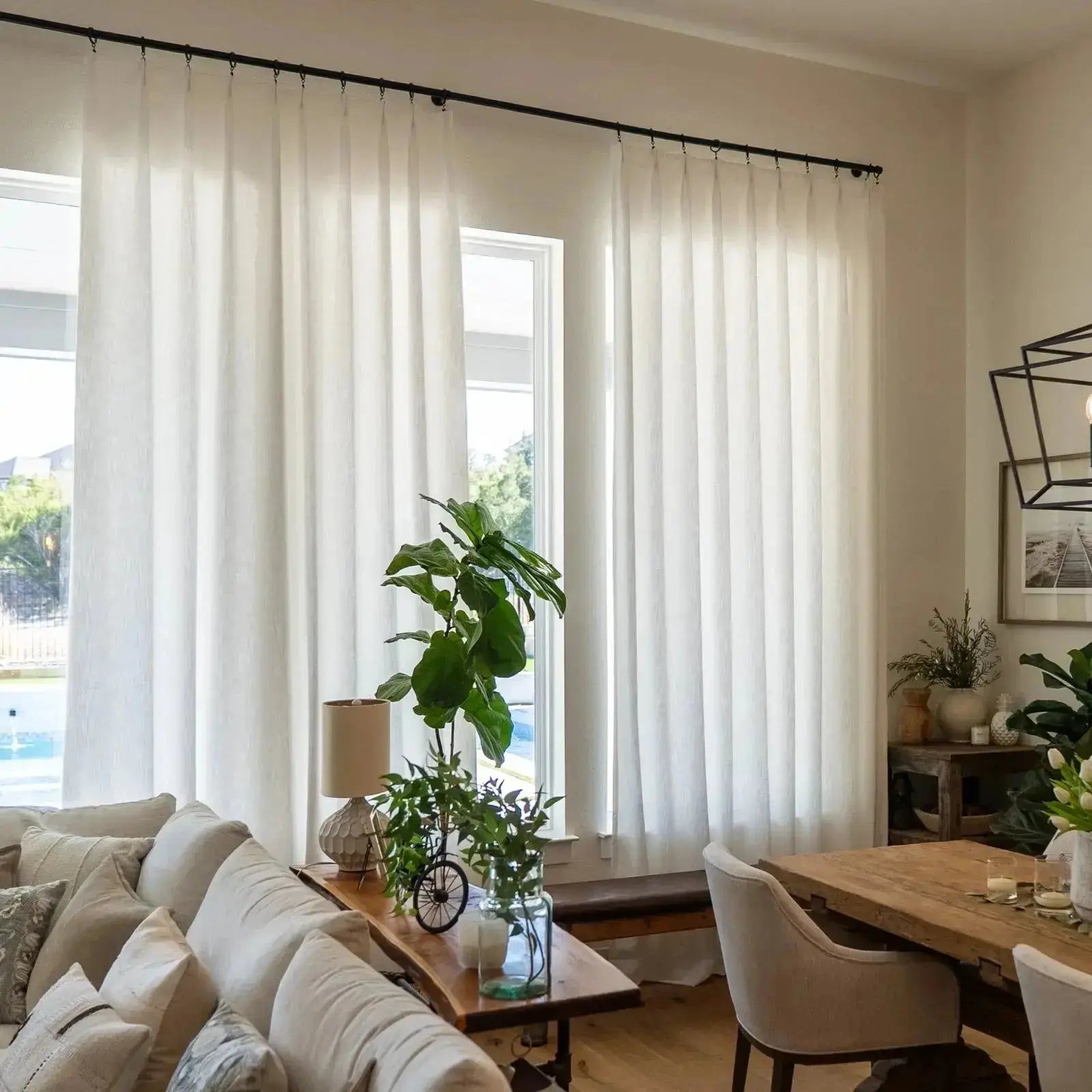 Interior view of a modern living dining room featuring sheer white linen curtains a large fiddle-leaf fig plant a geometric black chandelier and a visible outdoor swimming pool