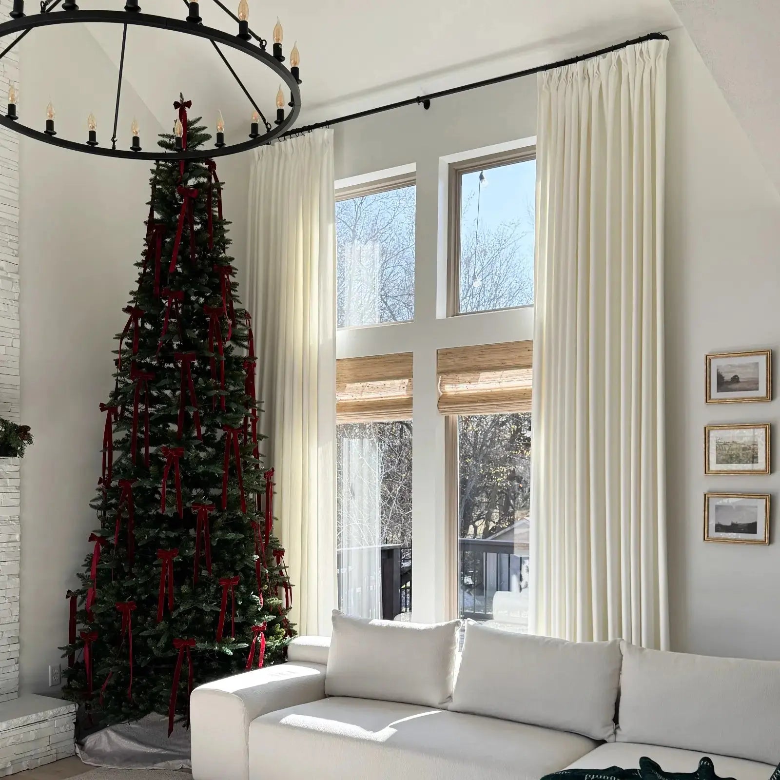 A modern living room features a tall Christmas tree adorned with red velvet bows, a large black circular chandelier, and a white modular sectional sofa bathed in natural light from expansive windows framed by cream drapes showing bare trees outside