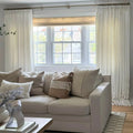 Photograph of a cozy modern living room interior featuring a light beige sofa with diverse neutral throw pillows, a large window overlooking a residential street, framed by white pleated floor-length curtains on a brass rod and a light purple floral arrangement in a ribbed white vase on the coffee table