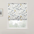 Photograph of a white Roman blind with an intricate pattern of birds perched on branches featuring yellow flowers and blue leaves, covering a window that looks out onto blurred greenery