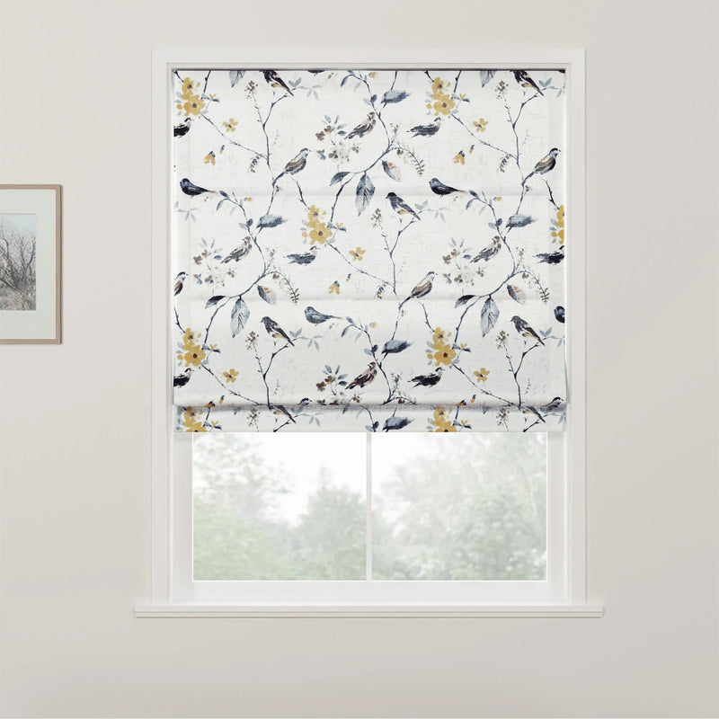 Photograph of a white Roman blind with an intricate pattern of birds perched on branches featuring yellow flowers and blue leaves, covering a window that looks out onto blurred greenery