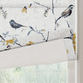 Fabric blind with a delicate bird and branch pattern featuring grey-blue leaves and vibrant mustard yellow flowers on a textured white background partially rolled down over a window with blurred green foliage