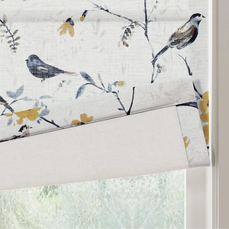 Fabric blind with a delicate bird and branch pattern featuring grey-blue leaves and vibrant mustard yellow flowers on a textured white background partially rolled down over a window with blurred green foliage