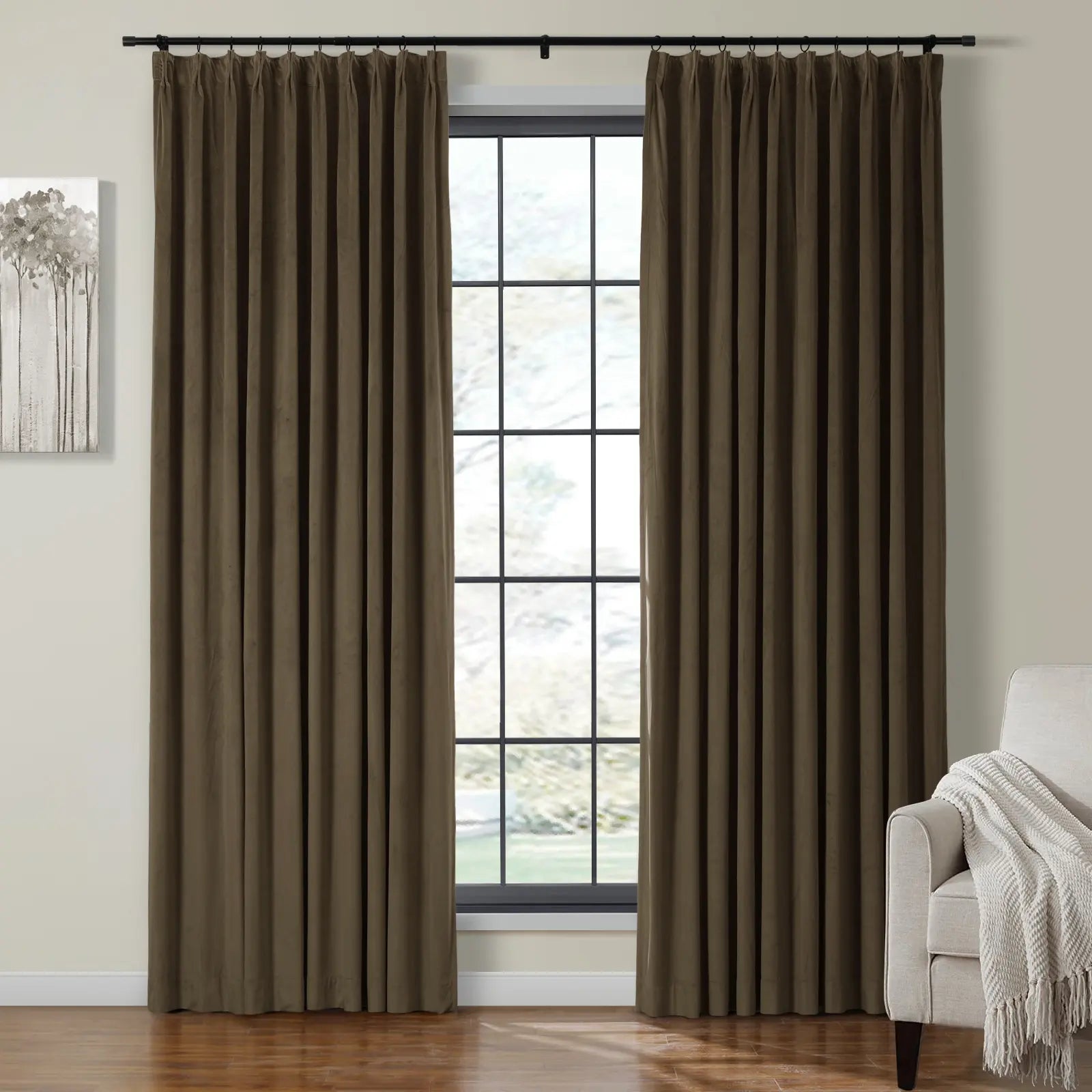 Dark brown pleated blackout curtains on a black rod frame a multi-pane window with a blurred green outdoor view in a modern interior featuring a cream armchair with a white woven throw and a gray abstract tree painting