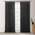 Charcoal black pinch-pleat curtains on a black rod frame a multi-pane window with diffused light and outdoor greenery, accompanied by a wooden fluted side table with a black vase and brass candlestick