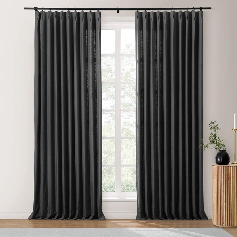 Charcoal black pinch-pleat curtains on a black rod frame a multi-pane window with diffused light and outdoor greenery, accompanied by a wooden fluted side table with a black vase and brass candlestick