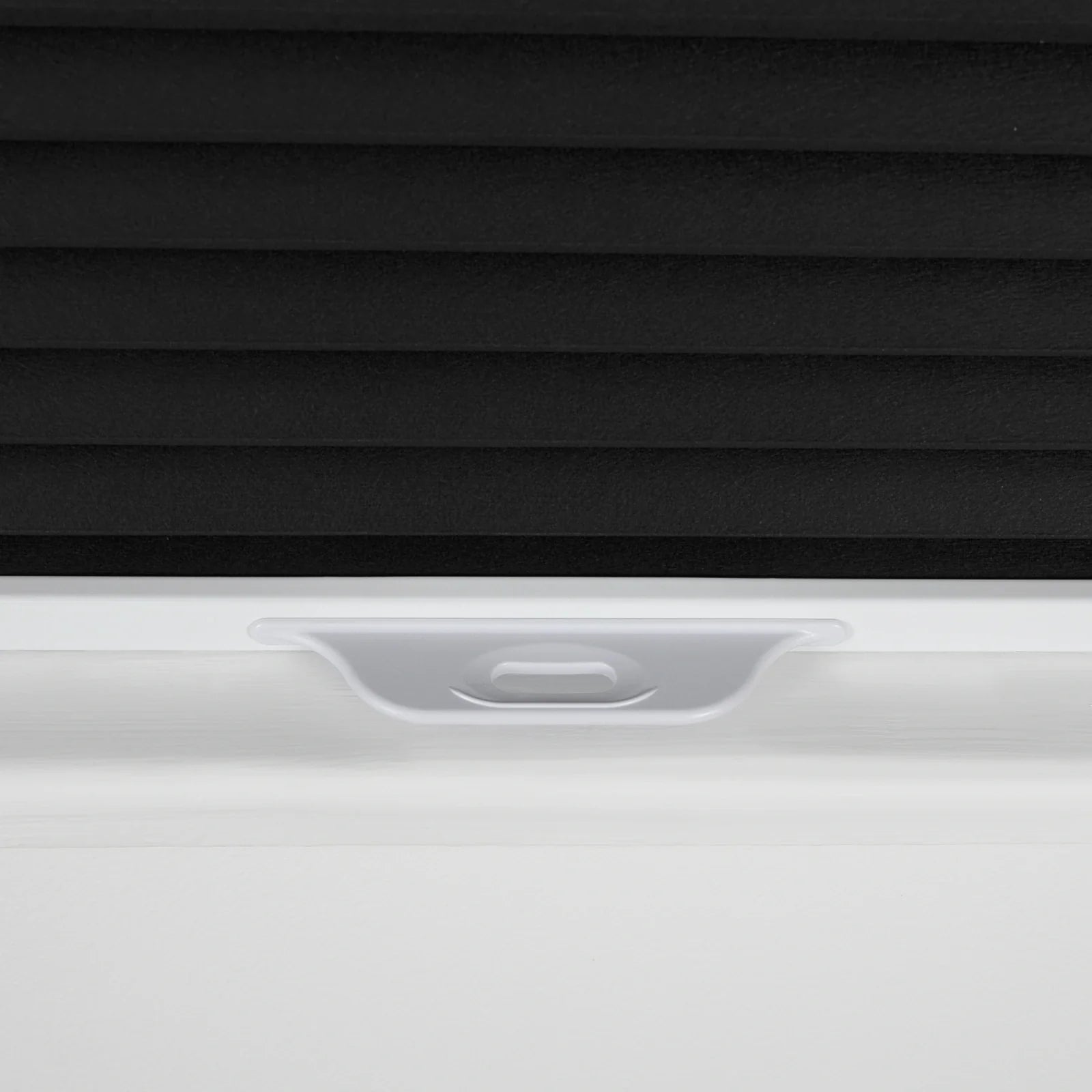 Product photography of a close-up detail of black textured cellular window Blinds partially lowered in a white window frame with a white plastic pull tab handle