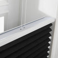 Close-up of a textured black pleated window Blind featuring a white headrail and two white control cords, set within a bright white window frame