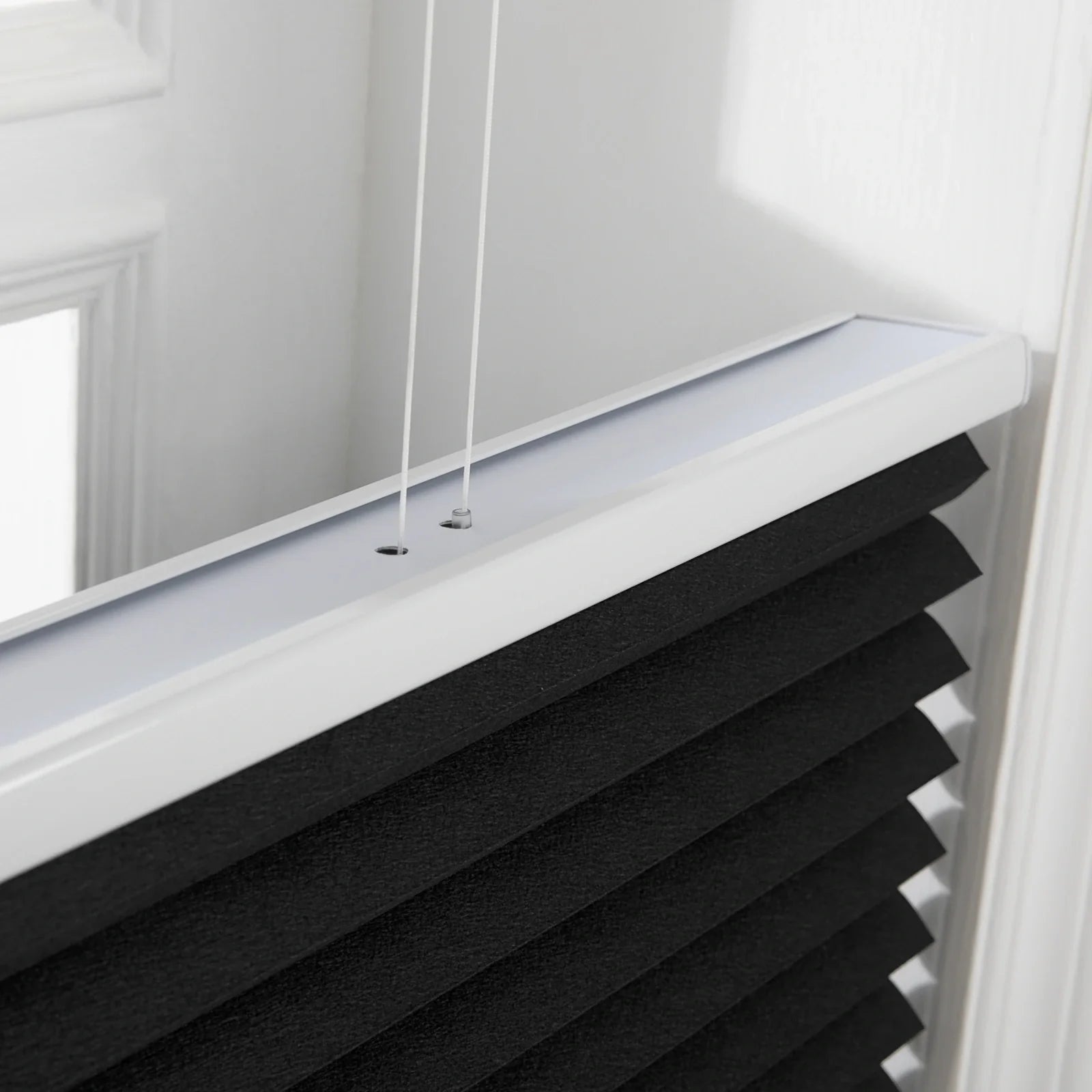 Close-up of a textured black pleated window Blind featuring a white headrail and two white control cords, set within a bright white window frame