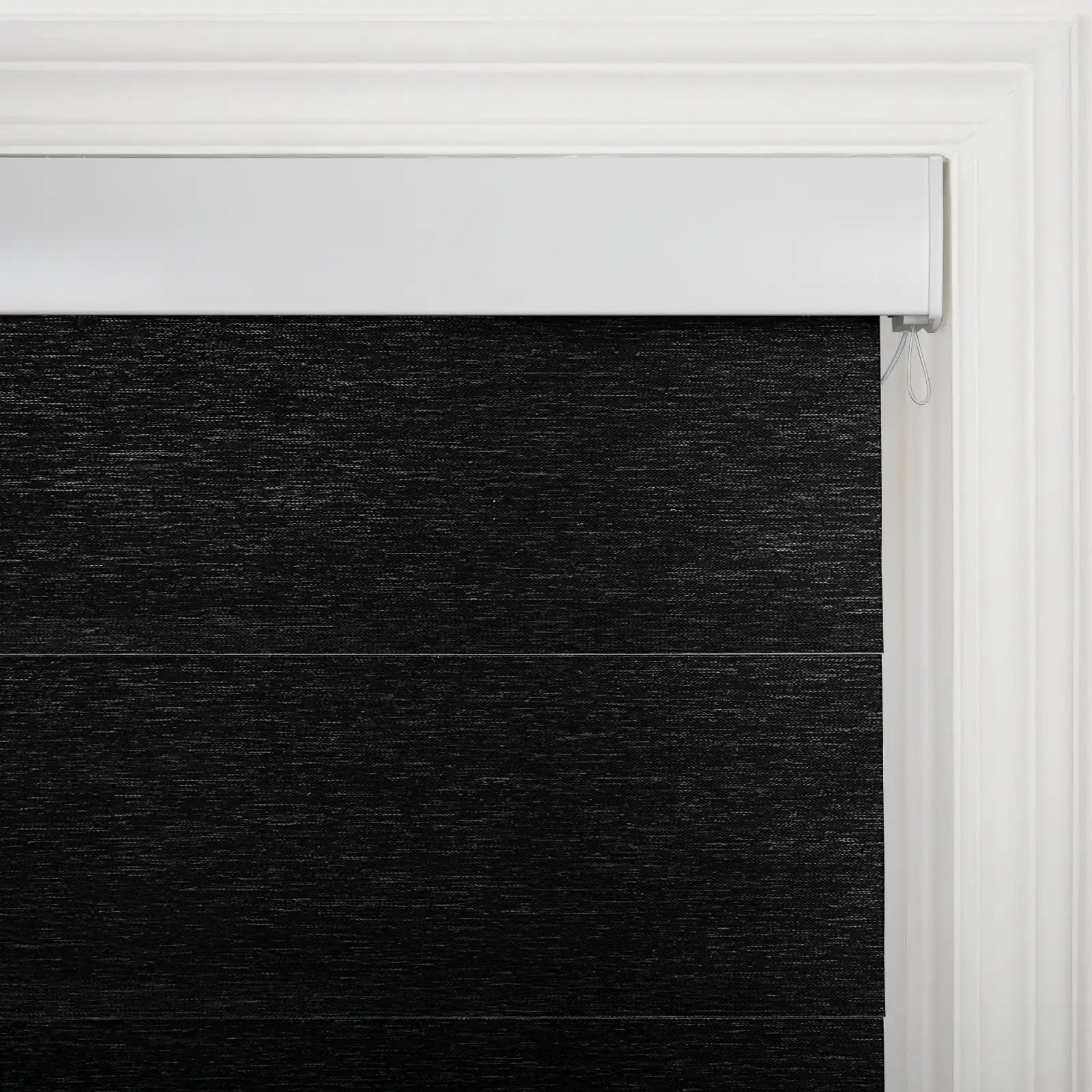 Close-up of a black textured fabric layered window Blind with a white headrail and a classic white molded window frame in an interior setting revealing the control cord