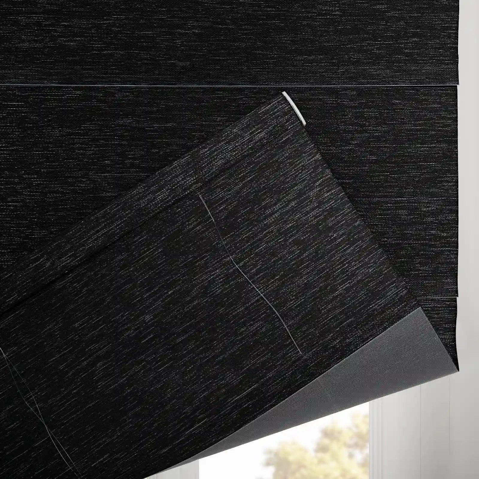 A close-up of a partially lowered black textured window Blind at a window, showing its subtle horizontal weave, grey thermal backing, and thin white lift cords with sunlight filtering from below