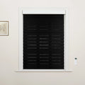 Photograph of a window fitted with black horizontal zebra Blinds showcasing alternating opaque and sheer fabric stripes, bordered by a crisp white frame, with a small white remote control on the right wall and a framed botanical print on the left