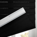 Black fabric zebra window Blinds with textured horizontal stripes partially lowered, showing the white headrail and a soft outdoor light from below
