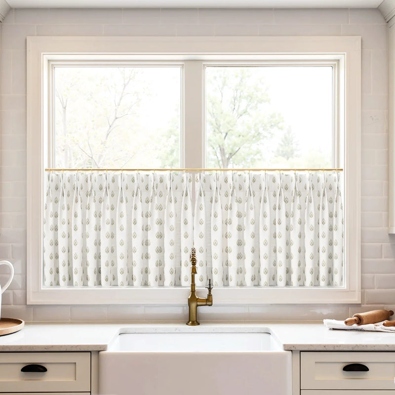 A kitchen window with a white cafe curtain featuring a golden teardrop pattern above a white farmhouse sink with a brass faucet and subway tile backsplash