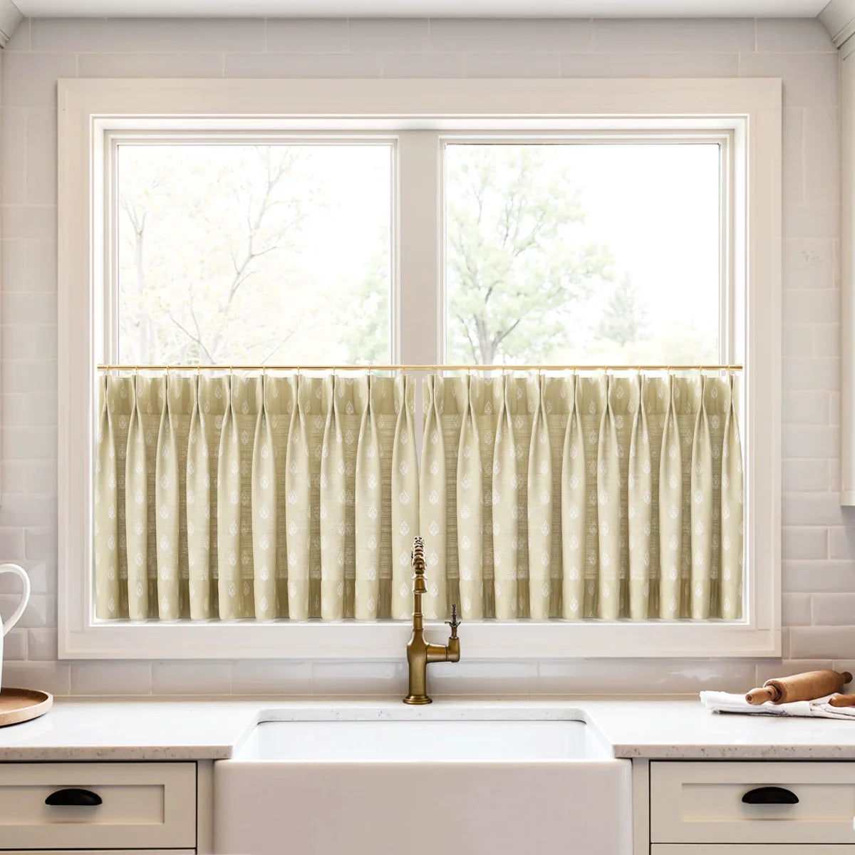 A kitchen window featuring pleated beige cafe curtains with a white leaf pattern, an antique brass faucet, and a white farmhouse sink