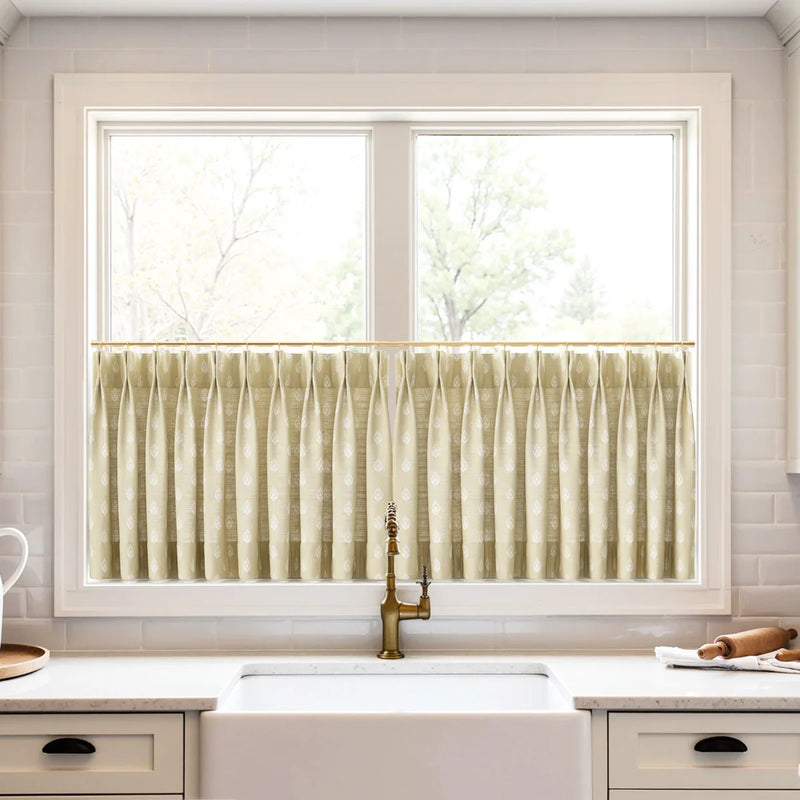 A kitchen window featuring pleated beige cafe curtains with a white leaf pattern, an antique brass faucet, and a white farmhouse sink