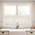 A modern kitchen window with white cafe curtains displaying a gold teardrop pattern centered above a white farmhouse sink and a distinctive brass faucet against a white subway tile backsplash