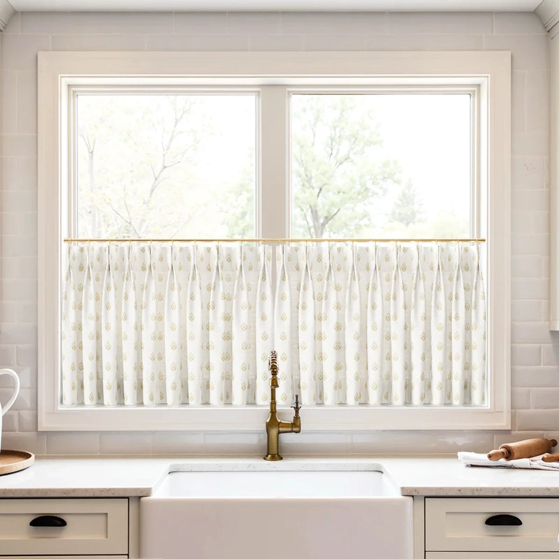 A modern kitchen window with white cafe curtains displaying a gold teardrop pattern centered above a white farmhouse sink and a distinctive brass faucet against a white subway tile backsplash