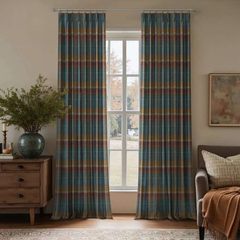 A cozy interior living space featuring a large window with teal gold and maroon plaid curtains a rustic wooden dresser adorned with a large green vase and foliage and a comfortable grey armchair with a cream patterned pillow and orange throw