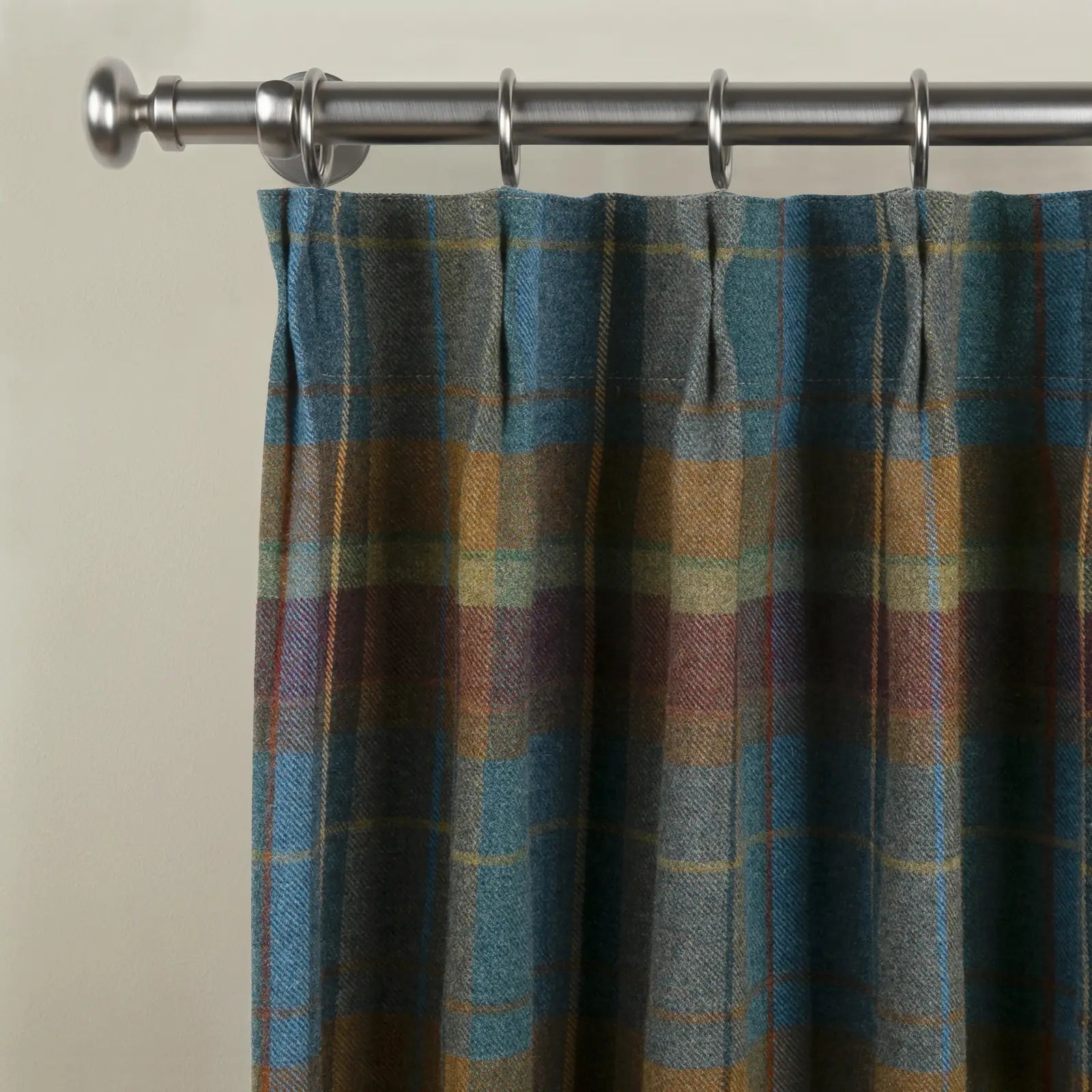 A rich wool plaid curtain in blue teal gold and earthy brown tones with pencil pleats hanging from a brushed nickel rod and rings