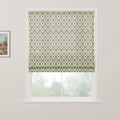 Product image of a Roman blind featuring a cream background with a repeating green leaf diamond pattern and small reddish-brown floral motifs partially drawn over a window revealing blurred green trees outside