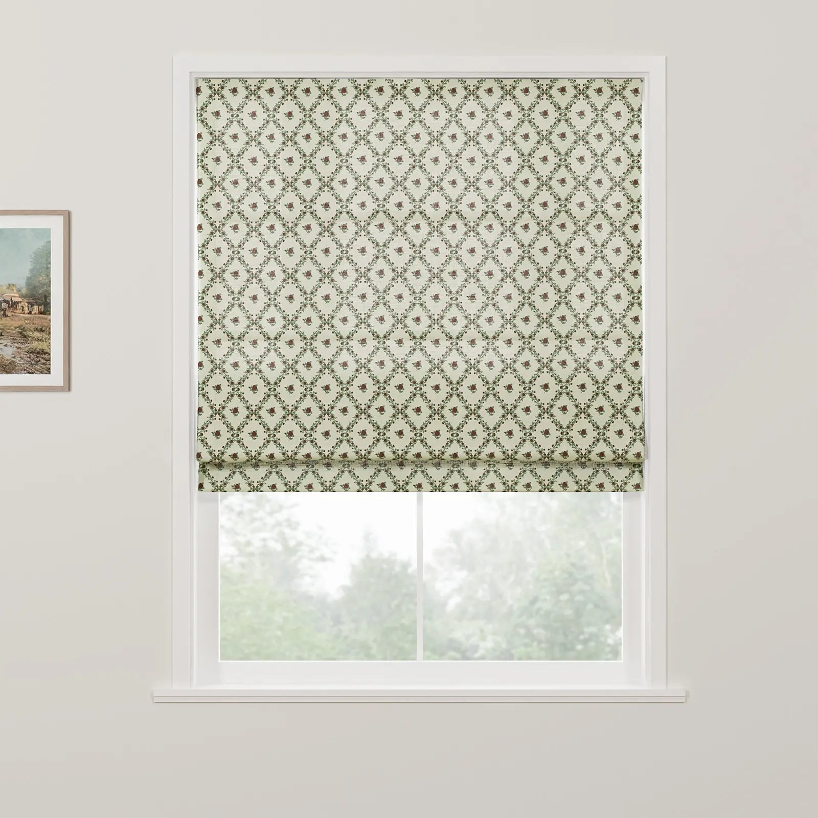 Product image of a Roman blind featuring a cream background with a repeating green leaf diamond pattern and small reddish-brown floral motifs partially drawn over a window revealing blurred green trees outside