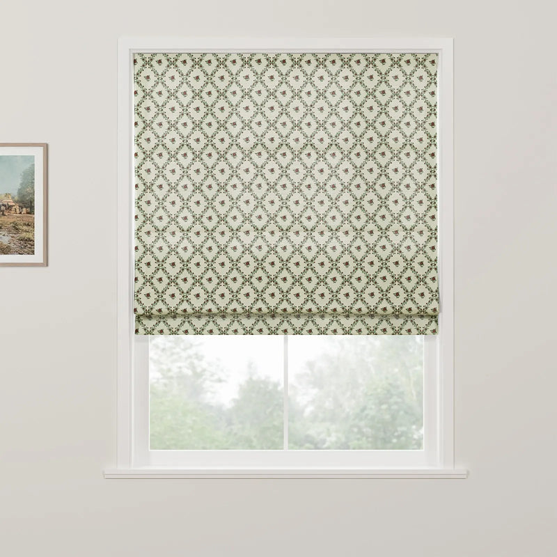 Product image of a Roman blind featuring a cream background with a repeating green leaf diamond pattern and small reddish-brown floral motifs partially drawn over a window revealing blurred green trees outside