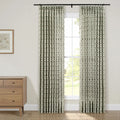 Pinch-pleated curtains in a white, green, and red geometric floral pattern hang from a black rod framing a window looking out to green foliage, alongside a natural wood dresser topped with a framed botanical print