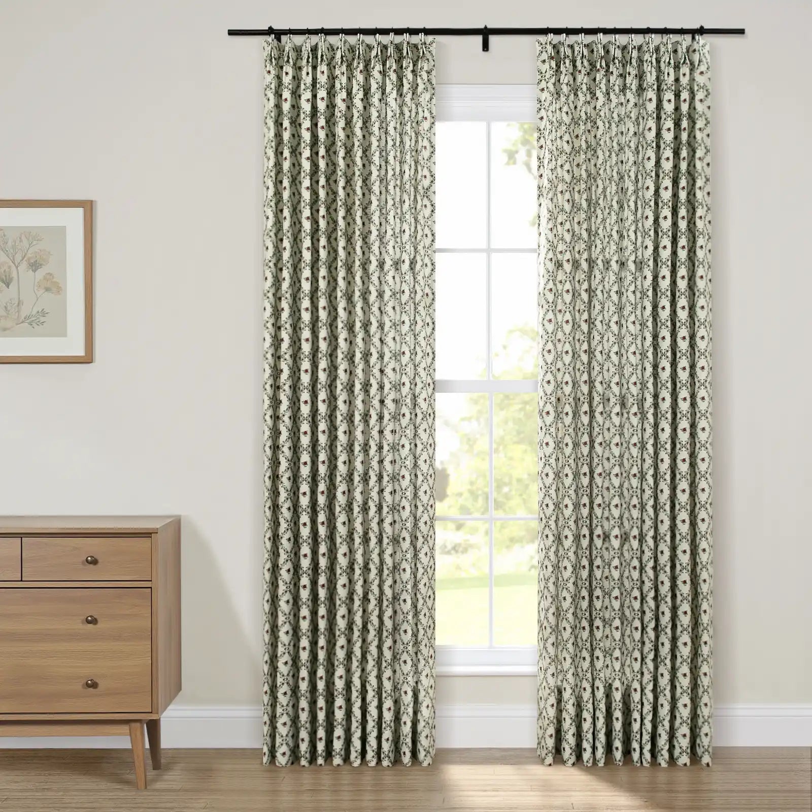 Pinch-pleated curtains in a white, green, and red geometric floral pattern hang from a black rod framing a window looking out to green foliage, alongside a natural wood dresser topped with a framed botanical print