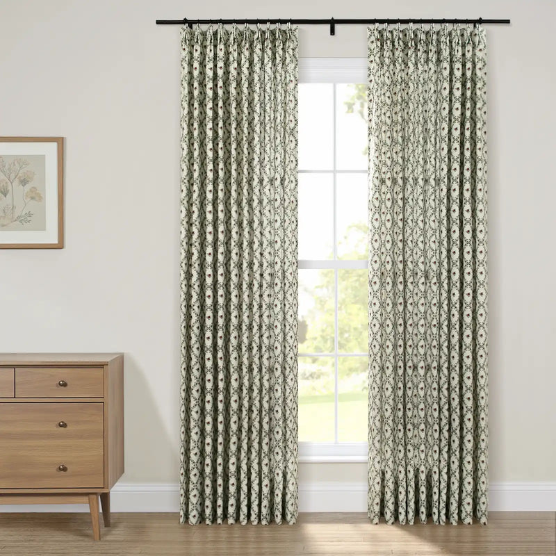 Pinch-pleated curtains in a white, green, and red geometric floral pattern hang from a black rod framing a window looking out to green foliage, alongside a natural wood dresser topped with a framed botanical print
