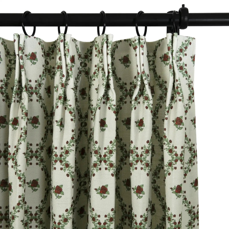 Close-up of an off-white pleated curtain with a repeating pattern of dark red roses and green leaves forming a diamond garland design hanging from a black rod with black rings