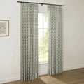 Interior room with light walls and a wooden floor showcasing pleated off-white curtains featuring a dark green diamond trellis pattern with small red floral motifs, hung from a black rod over a window, alongside a framed botanical print on the left wall