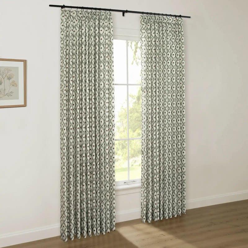 Interior room with light walls and a wooden floor showcasing pleated off-white curtains featuring a dark green diamond trellis pattern with small red floral motifs, hung from a black rod over a window, alongside a framed botanical print on the left wall