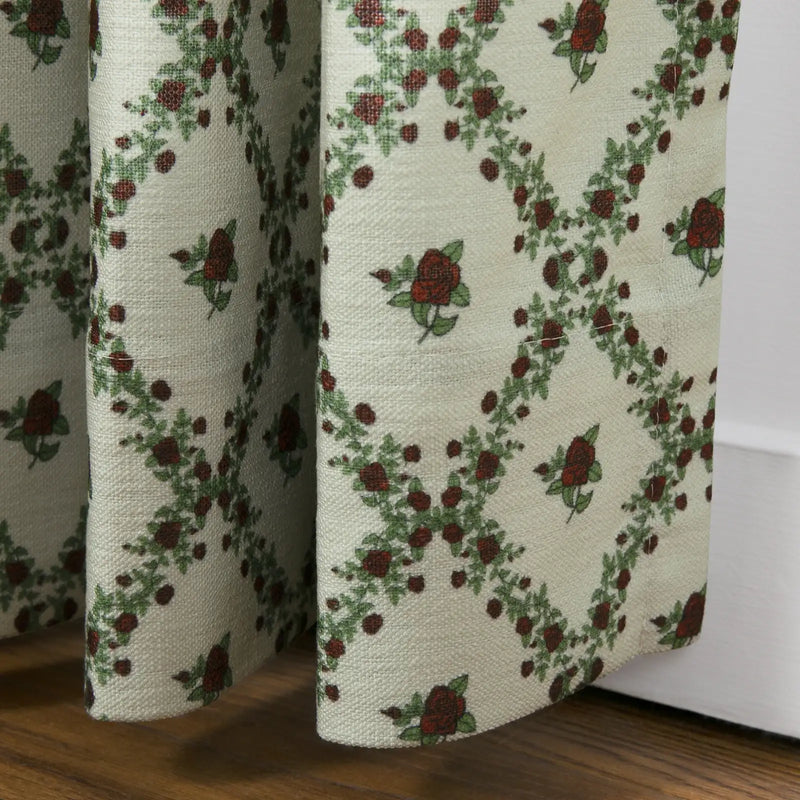 Textile: A cream-colored woven fabric curtain features a repeating trellis pattern of vibrant red roses and green leaves, hanging above a rich wooden floor beside a white baseboard