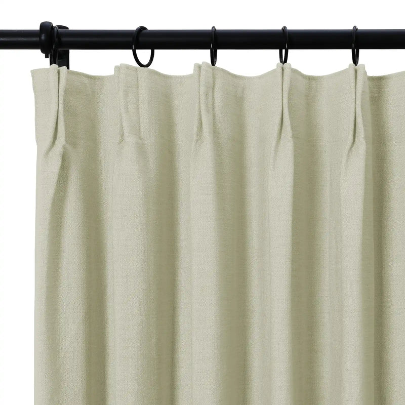 Photograph of a textured light oat-colored curtain with distinct pleats hanging from a black rod and matching rings