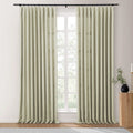 Light beige textured curtains on a black rod covering a grid window looking out to blurred greenery, with a fluted wooden side table, black vase, and gold candle holder on the right