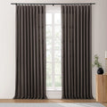 A pair of pleated dark gray linen-blend blackout curtains on a black rod frame a divided window with green trees outside, beside a fluted wooden table with a black vase of greenery and a tall brass candle holder