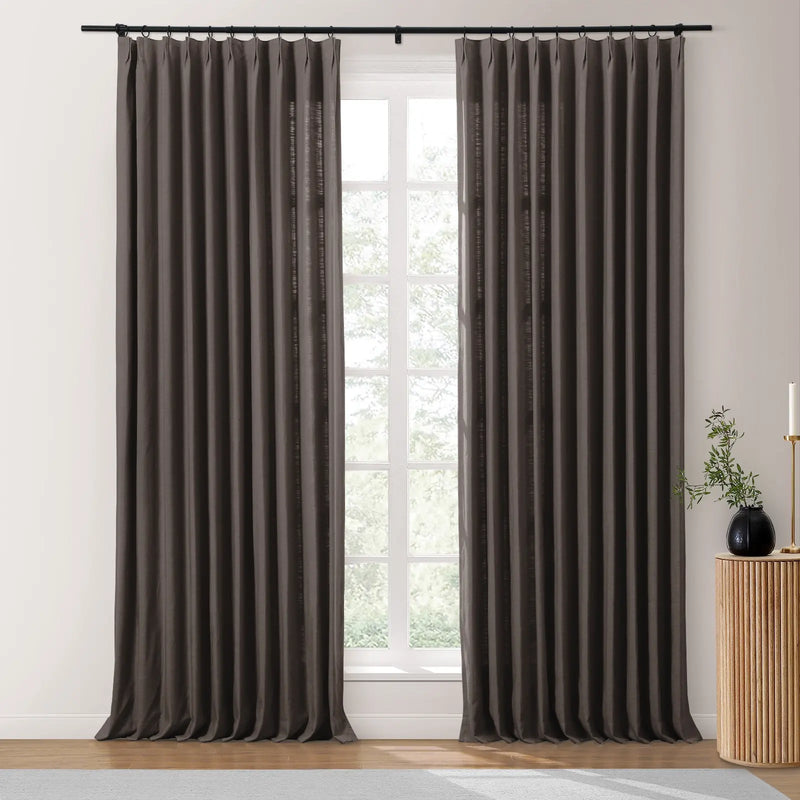 A pair of pleated dark gray linen-blend blackout curtains on a black rod frame a divided window with green trees outside, beside a fluted wooden table with a black vase of greenery and a tall brass candle holder