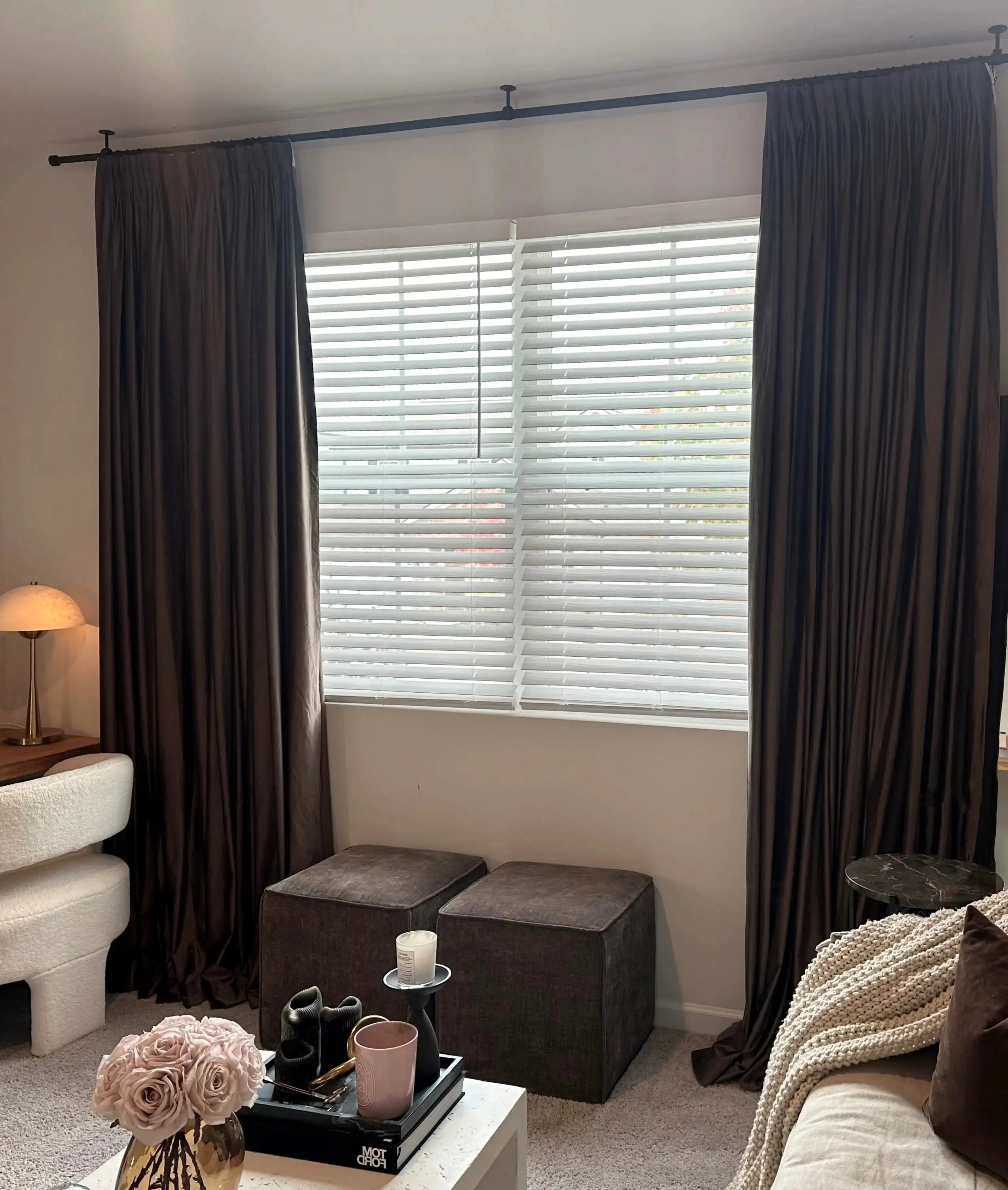 A contemporary living room interior featuring dark brown floor-to-ceiling drapes, white Venetian blinds, a plush white bouclé armchair, and light pink roses in a gold vase