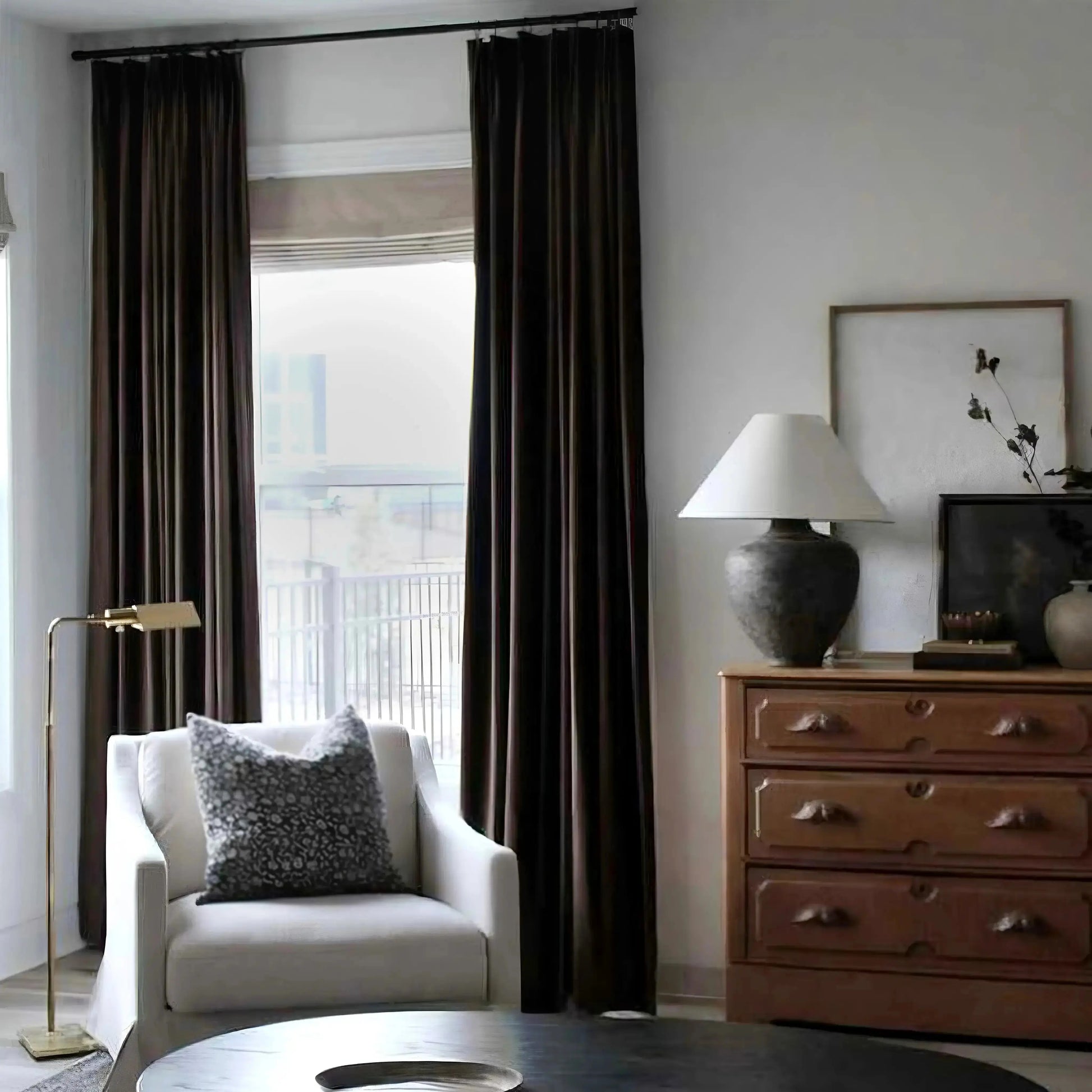 Photograph of a modern living room featuring rich dark brown velvet curtains framing a city view, a cream armchair with a black and white patterned pillow, and an ornate wooden dresser topped with a large grey lamp and dried botanicals