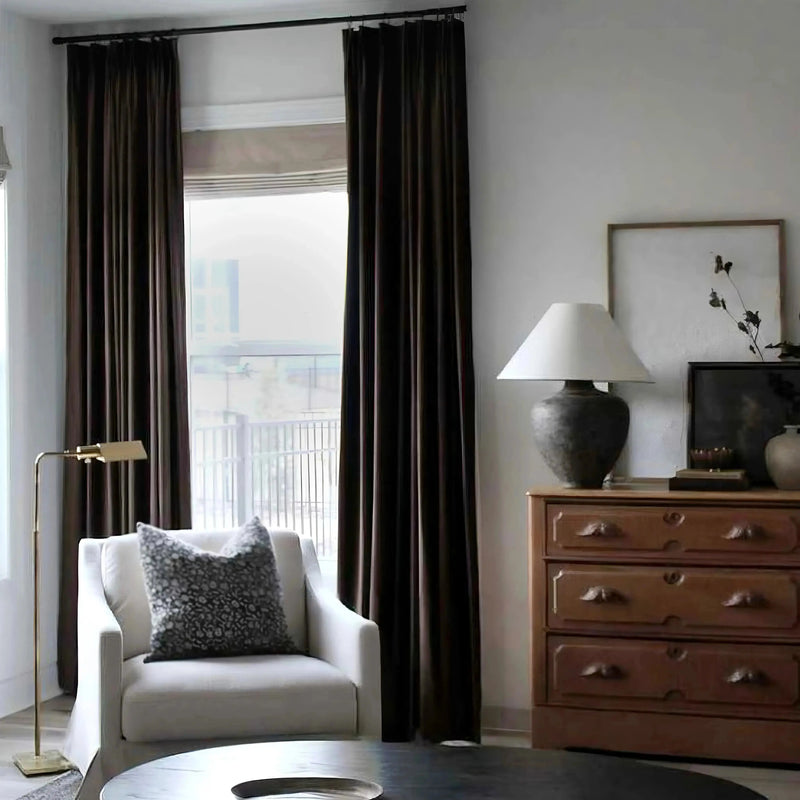 Photograph of a modern living room featuring rich dark brown velvet curtains framing a city view, a cream armchair with a black and white patterned pillow, and an ornate wooden dresser topped with a large grey lamp and dried botanicals